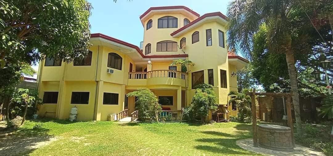 A spacious yellow house showcases multiple large windows and an expansive balcony surrounded by lush greenery. A well-maintained garden features various plants, and a traditional stone well is visible to the side, enhancing the tranquil outdoor setting.