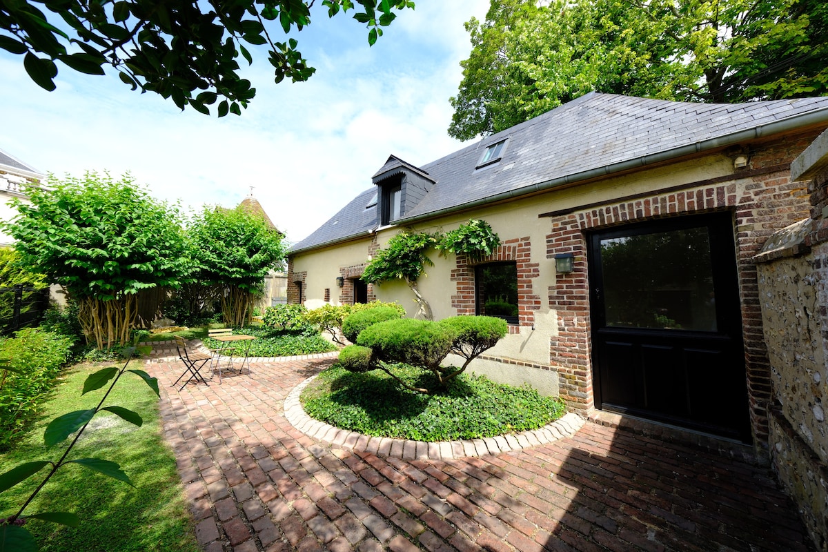 The exterior of the house is surrounded by a well-maintained garden featuring a circular brick pathway. Lush greenery and neatly trimmed shrubs enhance the inviting outdoor space. An entrance door is visible, along with two outdoor chairs positioned near the garden area.