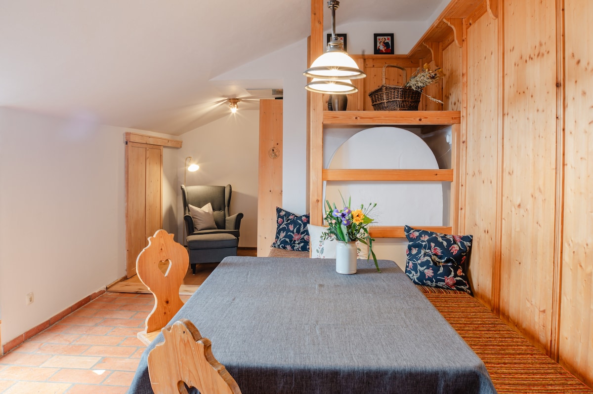 A dining area features a dark, tablecloth-covered table surrounded by wooden chairs. Natural wood paneling adorns the walls, and decorative cushions provide comfort. A softly lit corner includes a plush armchair, enhancing the cozy atmosphere of the space.