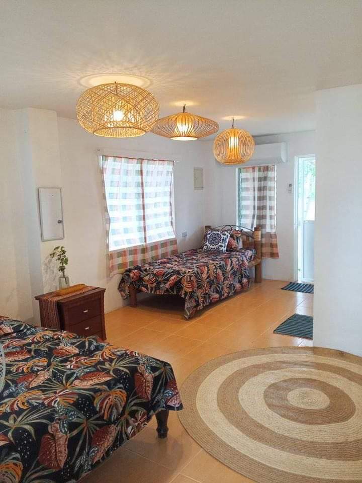 A bright and airy room is furnished with two beds, each adorned with patterned bedspreads in warm colors. Natural light filters through two windows dressed in checked curtains, while decorative woven lamps hang from the ceiling. A circular rug adds warmth to the floor.