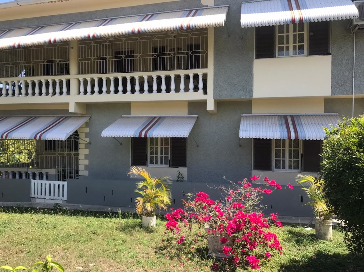 The exterior of a two-story house is displayed, featuring a light grey facade and decorative striped awnings over the windows. Lush greenery surrounds the house, with a vibrant pink flowering plant situated in front, adding a splash of color to the welcoming entrance.