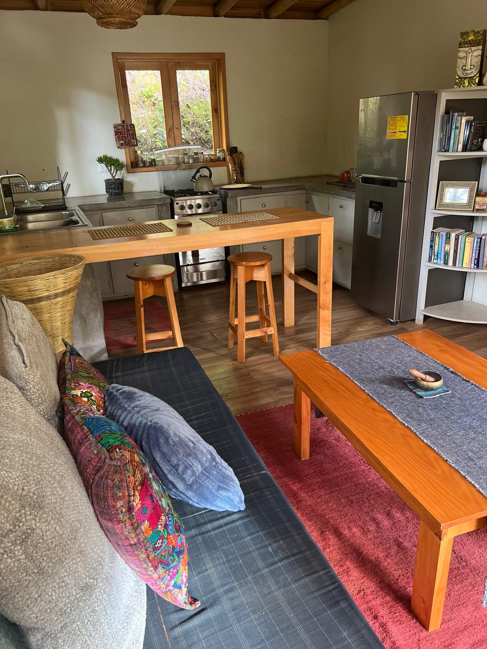 A cozy kitchen and living area features a wooden table with two stools. A silver refrigerator and a bookshelf filled with books are visible. Soft cushions in various colors are arranged on a gray sofa, complementing a wooden coffee table at the center.