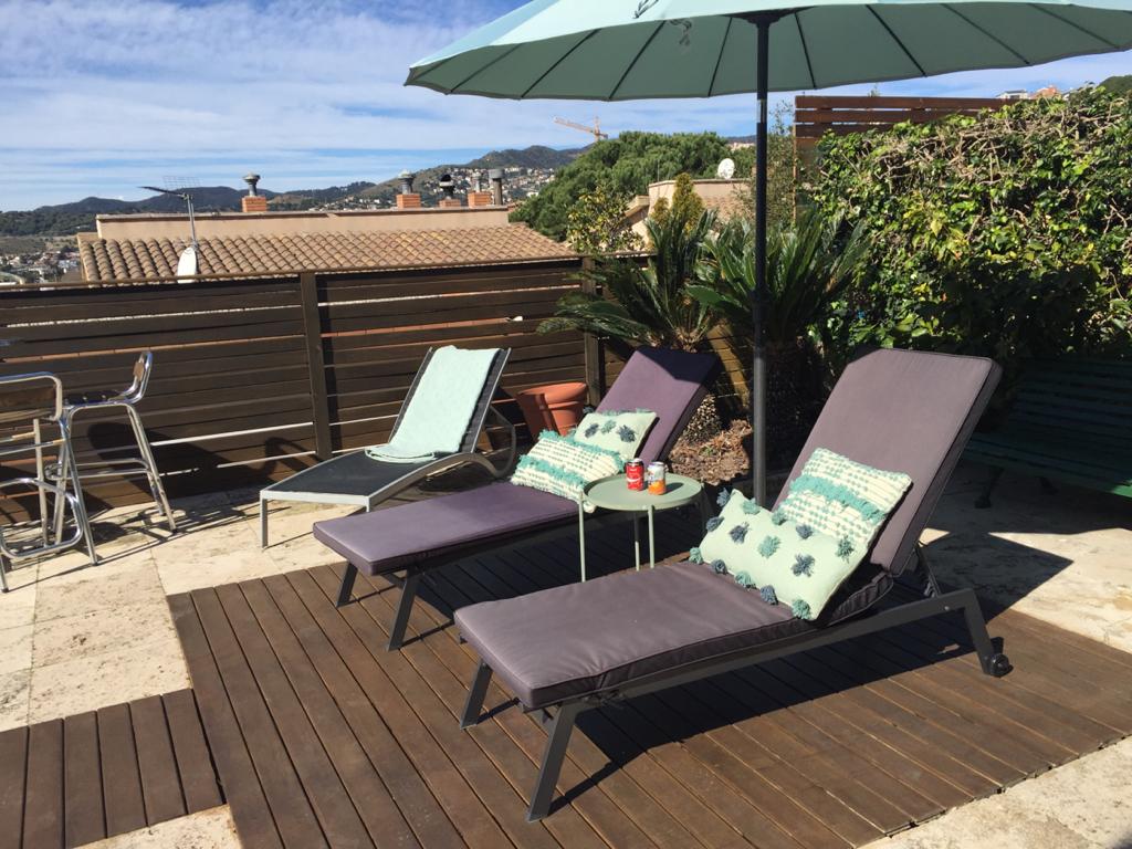 Two lounge chairs are positioned on a wooden deck, complemented by a small table featuring drinks. An umbrella provides shade, while surrounding greenery enhances the outdoor space. Scenic views of rolling hills can be seen in the background under a clear blue sky.