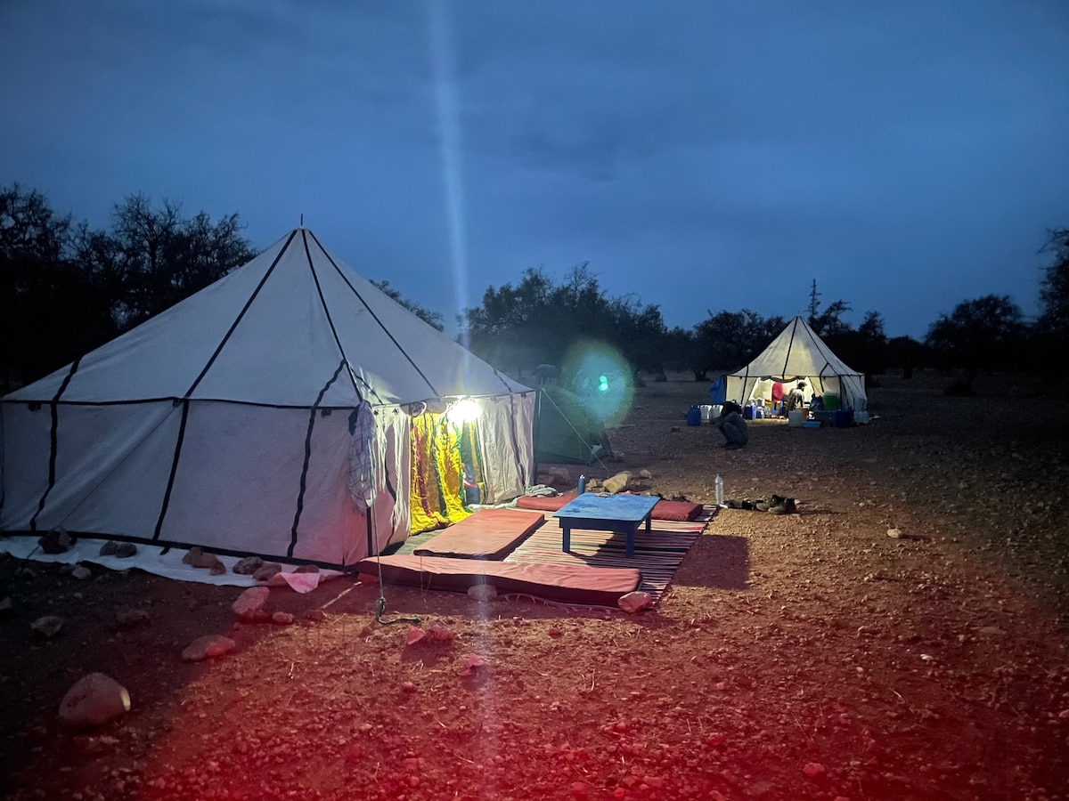A large, traditionally styled tent is illuminated by warm light, highlighting colorful decorations at the entrance. Nearby, another tent can be seen, set against a darkening sky. The ground is covered with rocky terrain, surrounded by sparse vegetation.