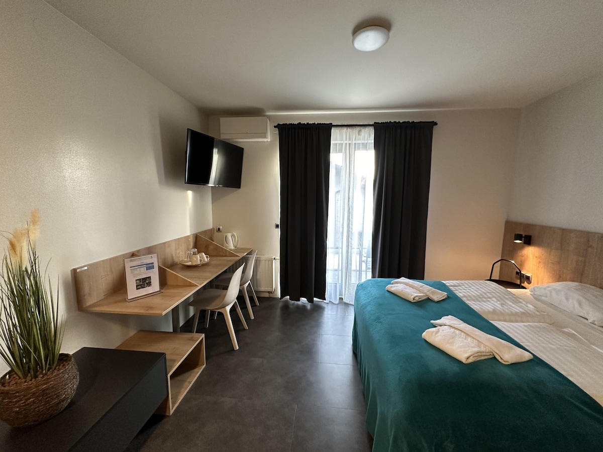 The room features a spacious bed with crisp linens and neatly arranged towels. A small table with two chairs is positioned next to a window draped with dark curtains. A flat-screen TV is mounted on the wall, and a natural plant adds a touch of decor.