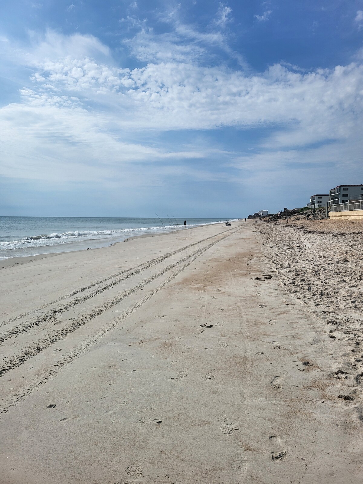 A serene beach scene is captured, featuring soft sand stretching alongside gentle waves. The shoreline shows tire tracks leading towards the distant buildings, under a clear sky with scattered clouds. The tranquil atmosphere hints at relaxation and offers ample space for walking or leisure activities.