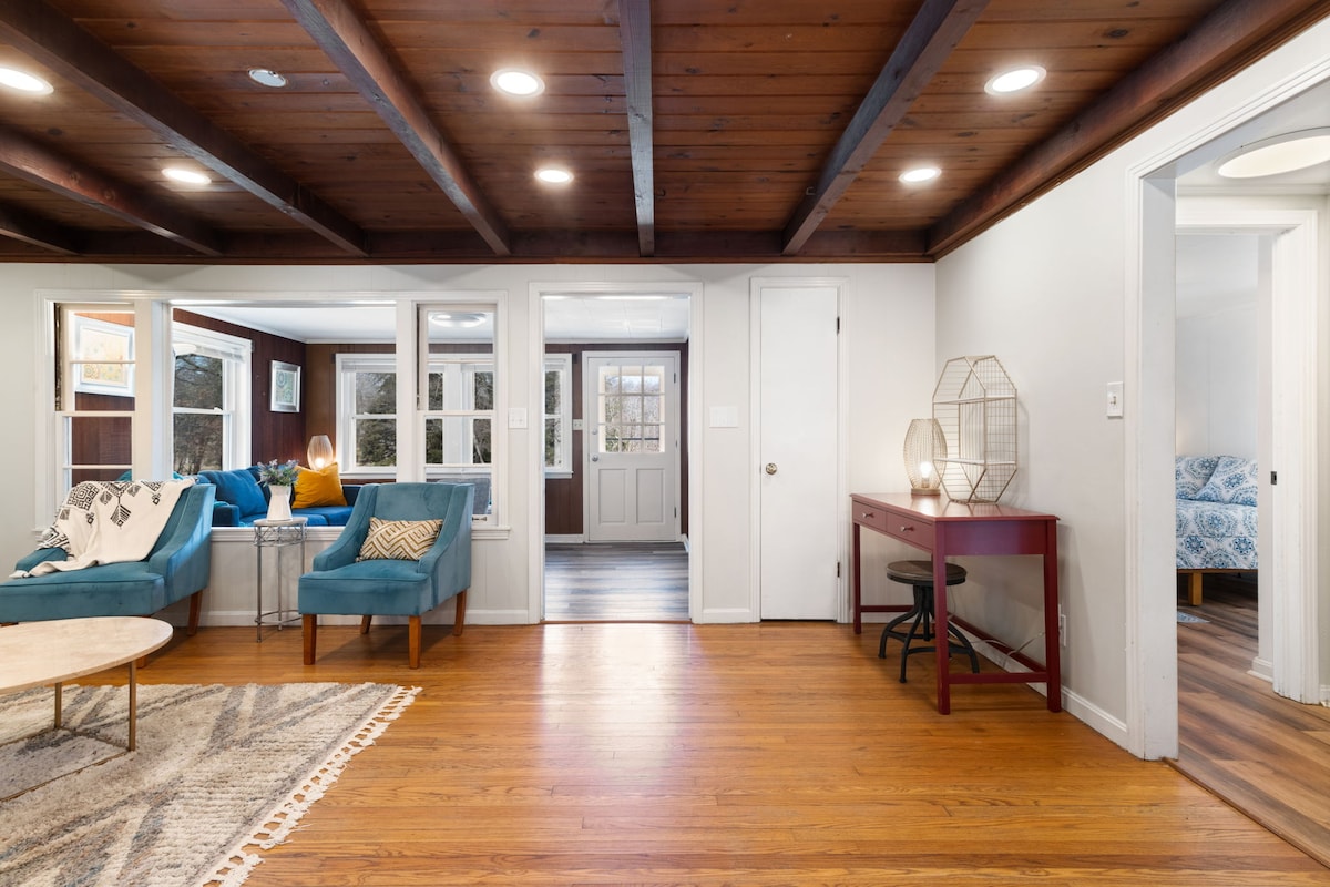 A spacious living area features comfortable turquoise seating and a round coffee table. Natural light streams through large windows, illuminating the wooden ceiling beams. A desk and chair are positioned against a wall, while an entrance leads to other rooms.