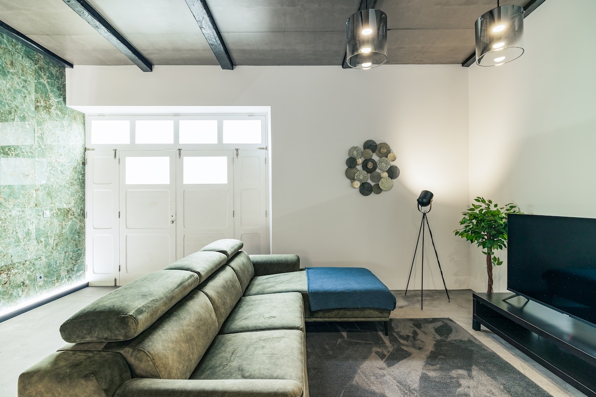 A spacious living area features a green sectional sofa with a blue accent, positioned next to a dark flat-screen television. Natural light floods the room through large windows. Decorative elements include a modern floor lamp and a wall-mounted art piece, enhancing the contemporary aesthetic.