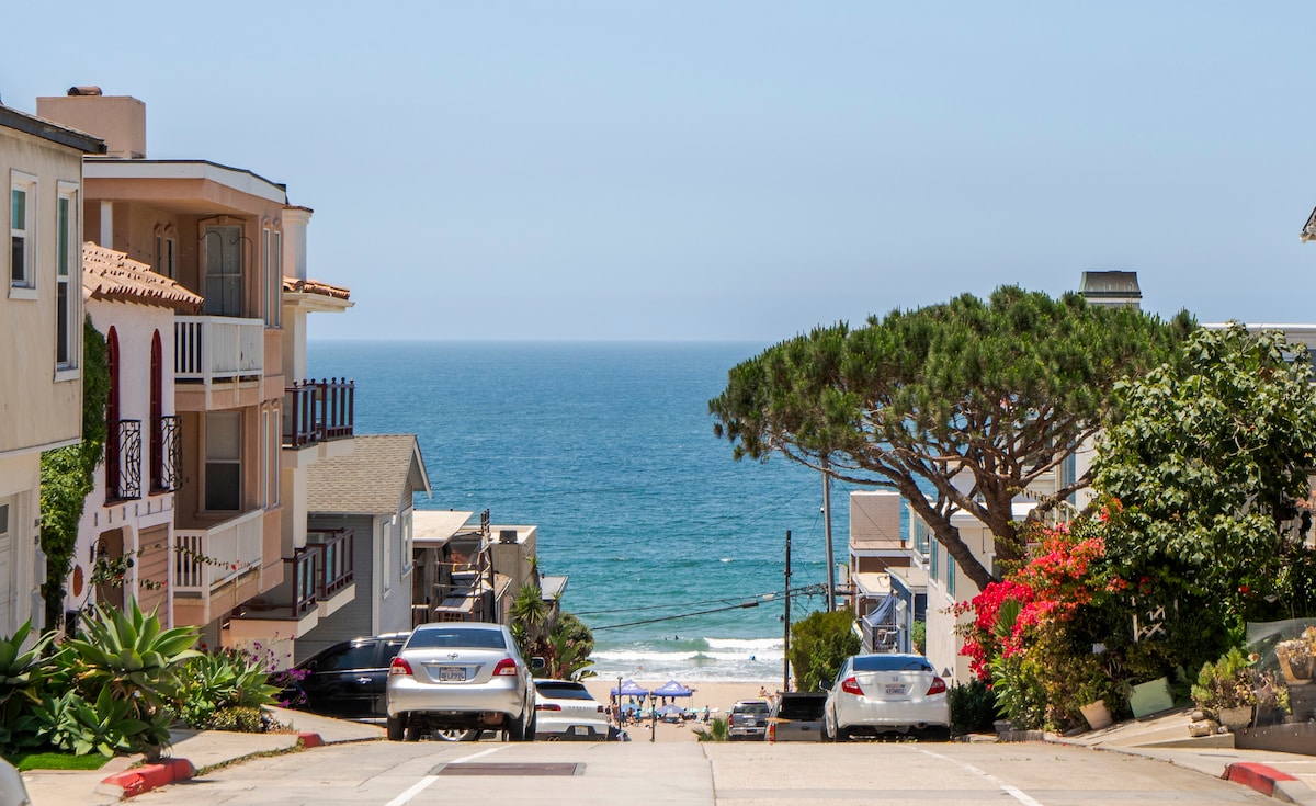 A serene coastal view features a sloping street leading down to the ocean. Vibrant flowers and greenery adorn the roadside, while distant beachgoers can be seen along the sandy shore. The sky is clear, revealing the calm blue water and gentle waves.