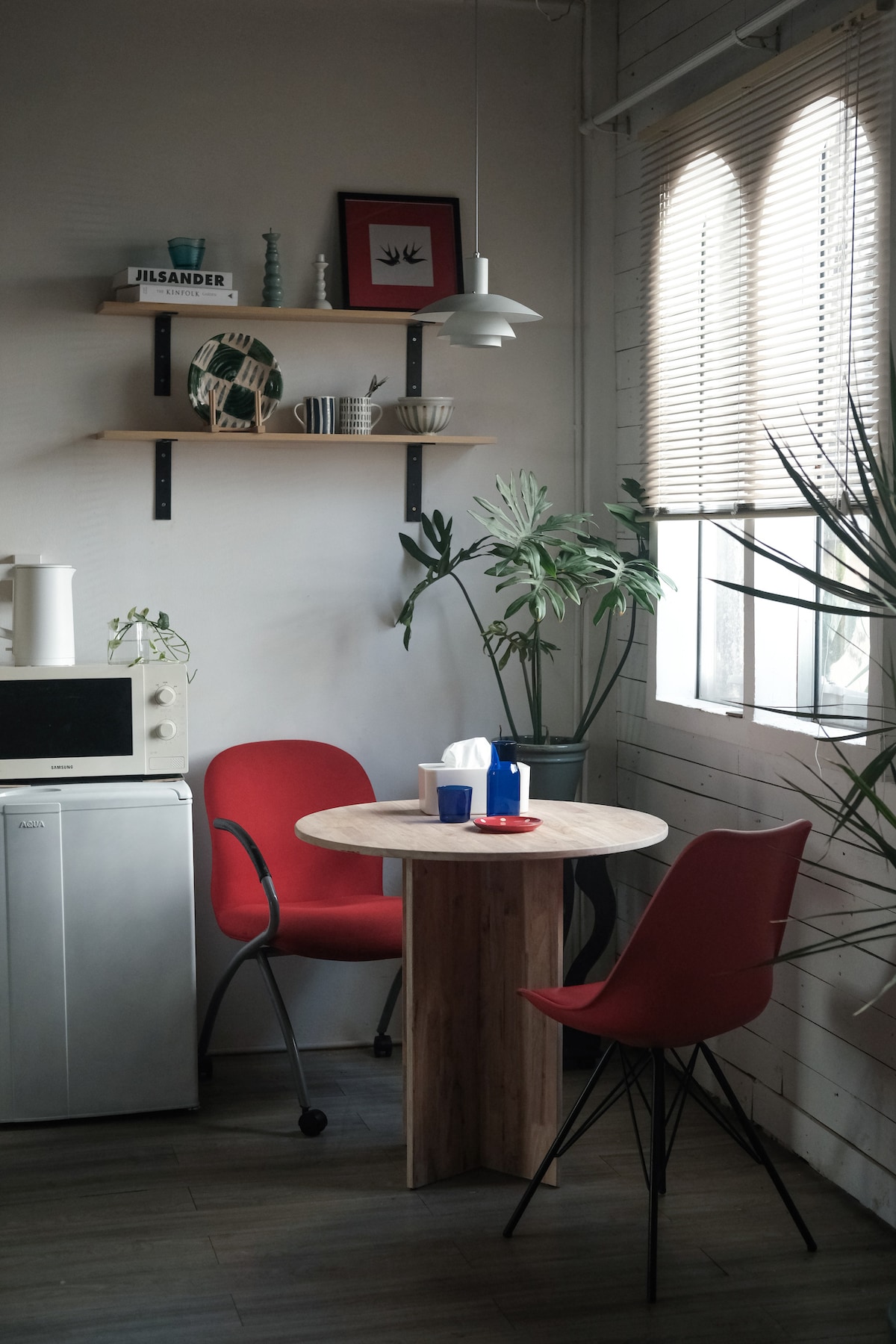 A cozy dining area is depicted with a round wooden table surrounded by two red chairs, one of which has wheels. A wall-mounted shelf displays decorative items alongside green plants. Natural light enters through an arched window, enhancing the simplicity of the space.