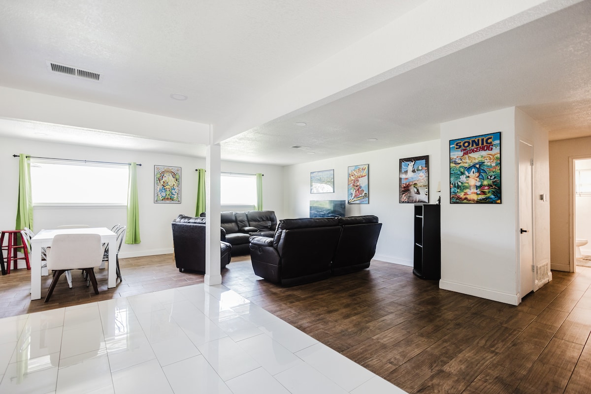 A spacious living area features light tile flooring and natural light entering through large windows with green curtains. Comfortable leather seating is arranged around a coffee table, while colorful wall art adds character. A dining table with chairs occupies one corner, promoting a welcoming environment.
