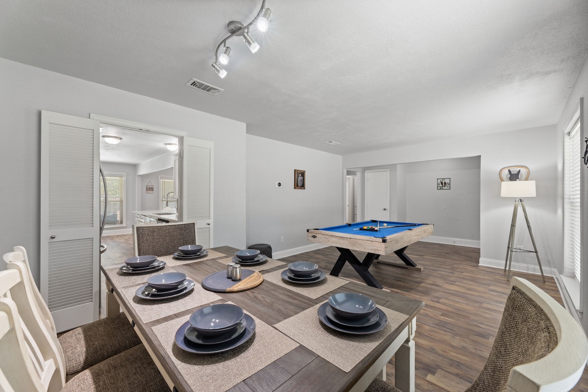 A dining area is paired with a modern pool table, providing options for both meals and entertainment. The long wooden table is set with dark plates, while neutral-colored seating surrounds it. Natural light illuminates the space, creating an open and inviting environment.