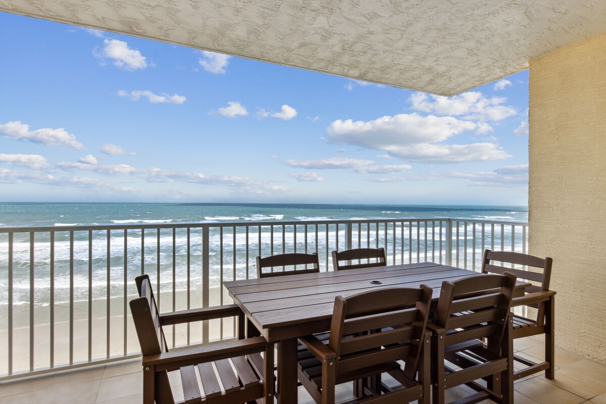A spacious outdoor balcony features a wooden dining table surrounded by matching chairs, with an unobstructed view of the ocean waves and blue sky. The railing provides safety while maintaining an open atmosphere, inviting guests to enjoy alfresco dining or leisurely moments at the coast.