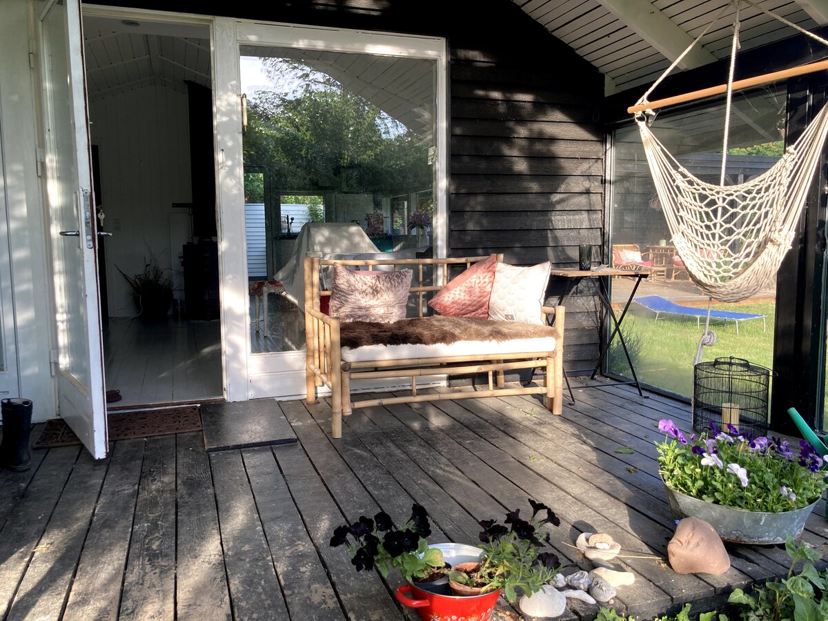 An outdoor seating area is presented with a wooden bench featuring cushions. A hammock is suspended nearby, and potted plants add greenery to the space. The entrance is framed by glass doors that open into the interior, creating an inviting transition.