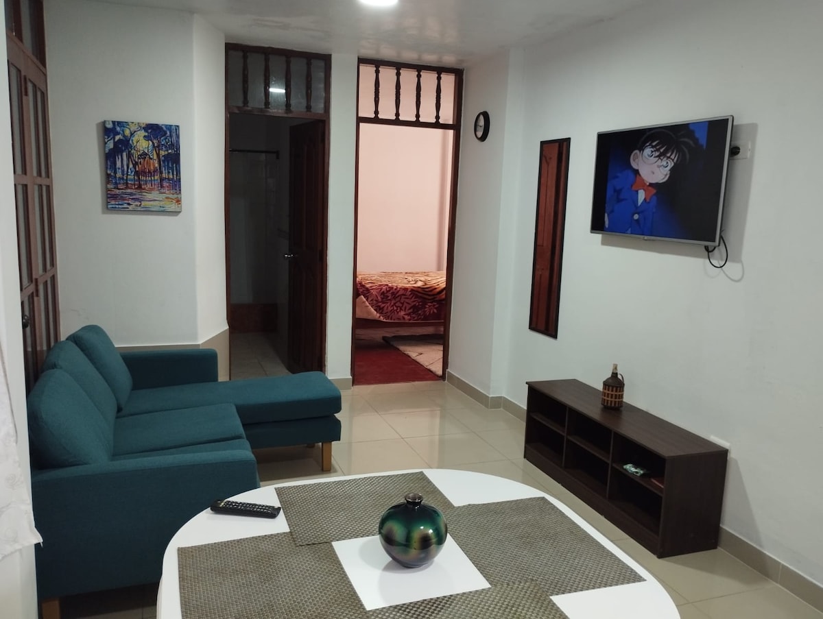 A well-arranged living area featuring a blue sofa and a round dining table. A television is mounted on the wall, and a decorative vase sits at the center of the table. A doorway leads to a bedroom in the background, with artwork displayed on the wall.