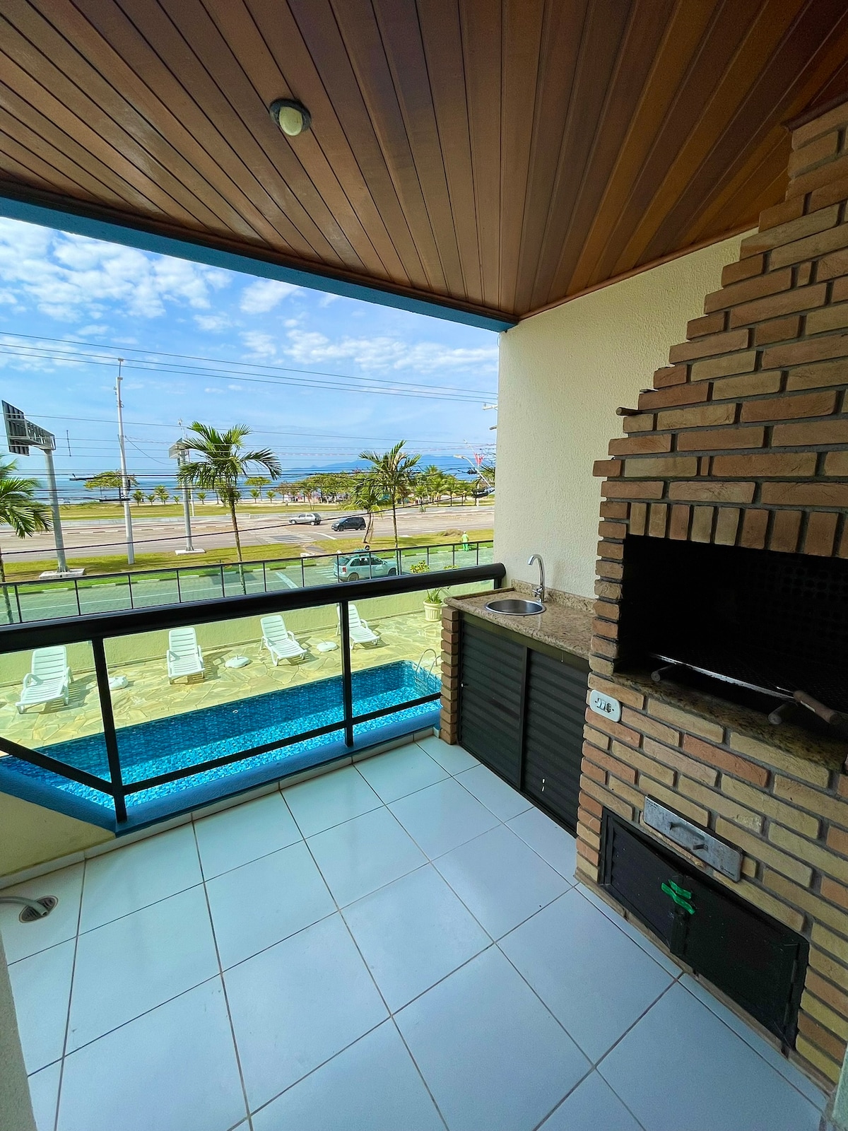 A private balcony features a built-in barbecue grill and a small sink, framed by a glass railing. The view includes a swimming pool and the ocean in the distance, with palm trees lining the area. The floor is tiled, and the ceiling showcases wooden paneling.