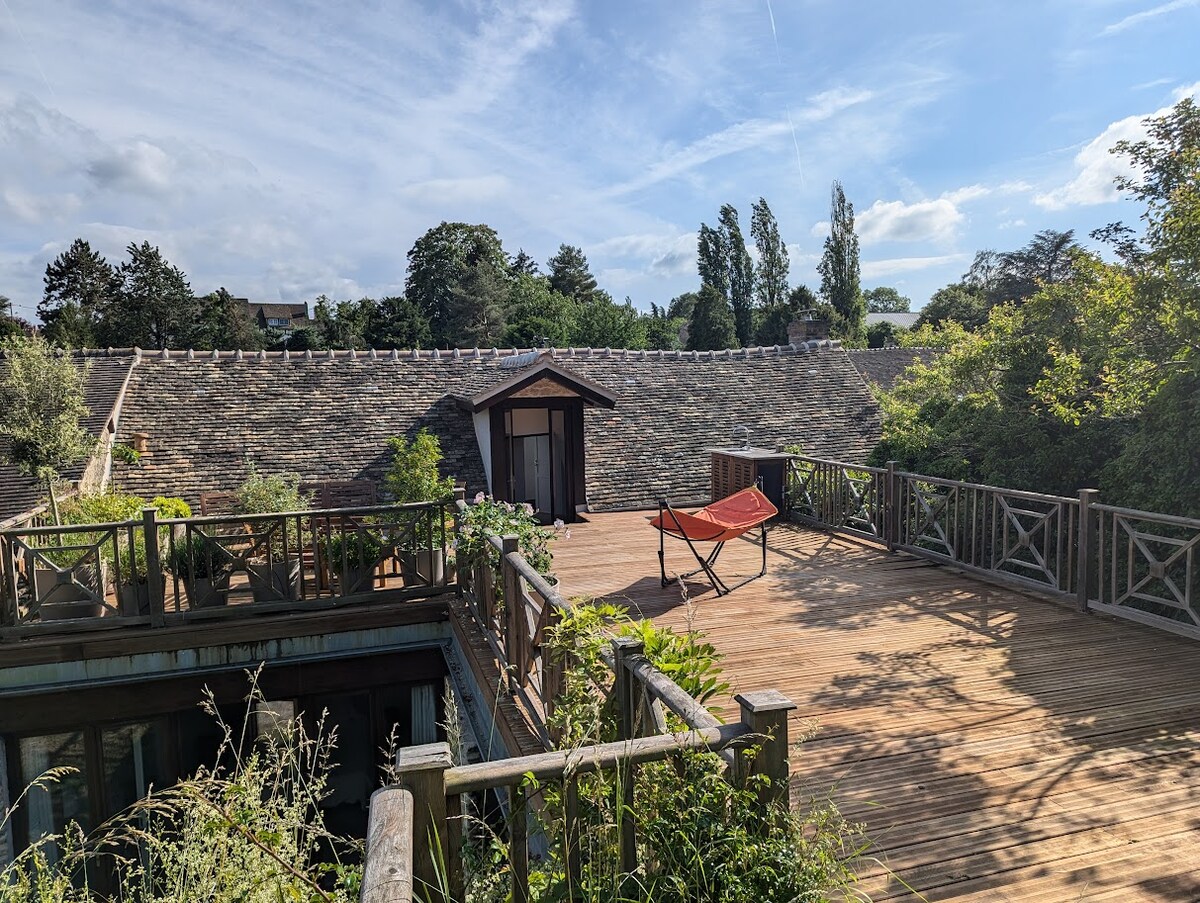 An expansive wooden deck surrounds a charming structure, highlighted by a small entrance. Lush greenery and plants are present, adding to the tranquil atmosphere. A hammock is positioned for relaxation, while the backdrop features a slate roof and a clear blue sky.