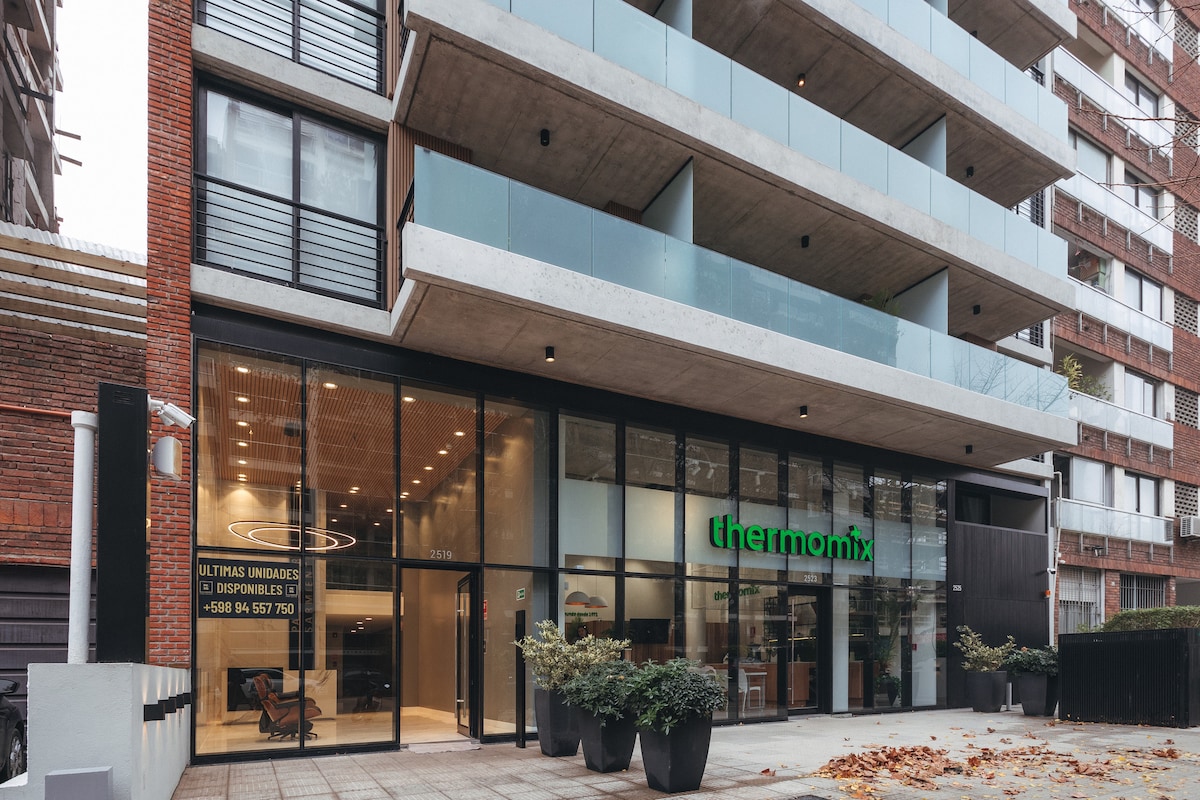 The modern building features a glass façade, allowing natural light to illuminate the interior. A green logo prominently displays the name 'thermomix.' Potted plants line the entrance, providing a welcoming touch to the urban setting.