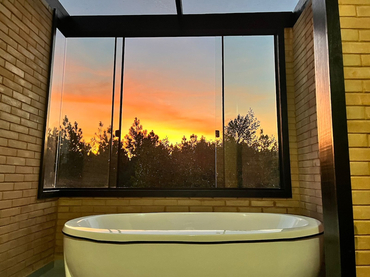 A spacious bathtub framed by large glass windows showcases a vibrant sunset view, blending warm hues with the silhouette of the surrounding trees. The light illuminates the room, accentuating the modern design featuring natural brick walls.