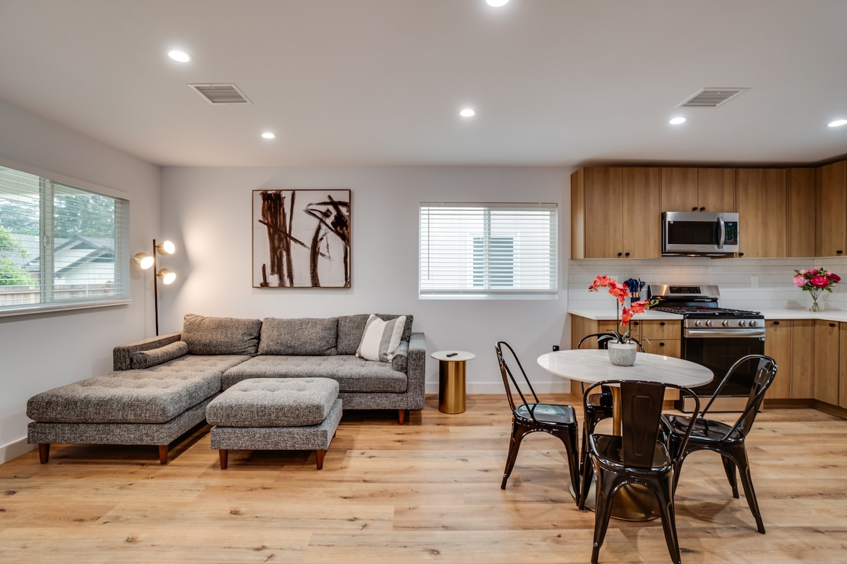 A modern living space is showcased with a gray sectional sofa and a dining area featuring a round table surrounded by four black chairs. Natural light floods in through two windows, illuminating the wood accents of the kitchen in the background.