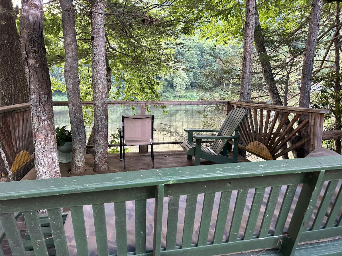 A shaded deck area is surrounded by trees, offering a peaceful view of the river. Comfortable seating options include a wooden Adirondack chair and a modern chair. The wooden railing complements the natural setting, inviting relaxation in a serene environment.
