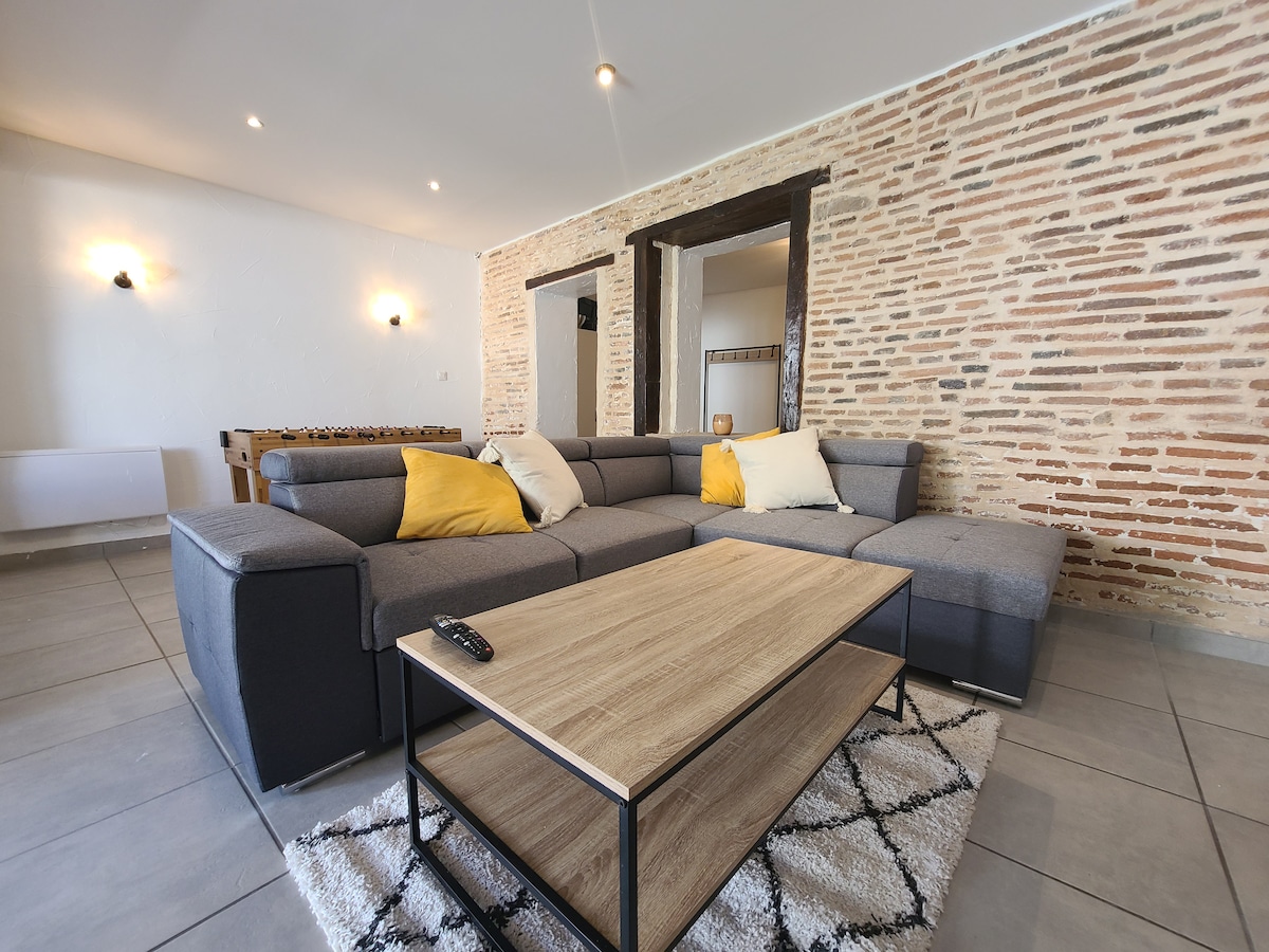 A spacious living area features a cozy sectional sofa, accented with yellow pillows. A wooden coffee table sits centrally on a textured rug, with a game table partially visible in the background. Light reflects off the exposed brick wall, adding warmth to the ambiance.