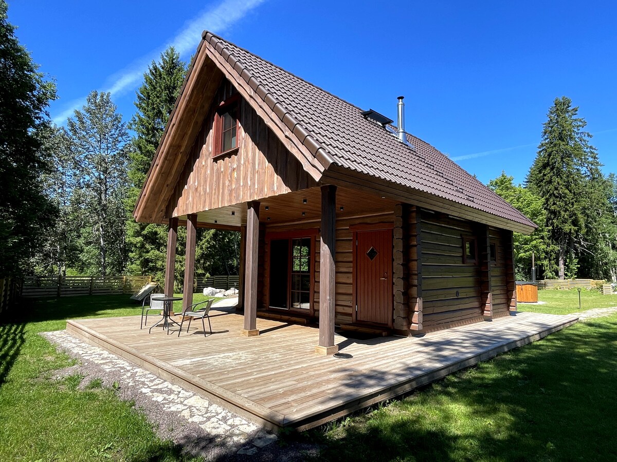 An inviting wooden cabin is set against a clear blue sky, featuring a sloped roof and spacious wooden deck. Two outdoor chairs are positioned on the deck, surrounded by greenery, while large windows allow natural light to fill the interior.