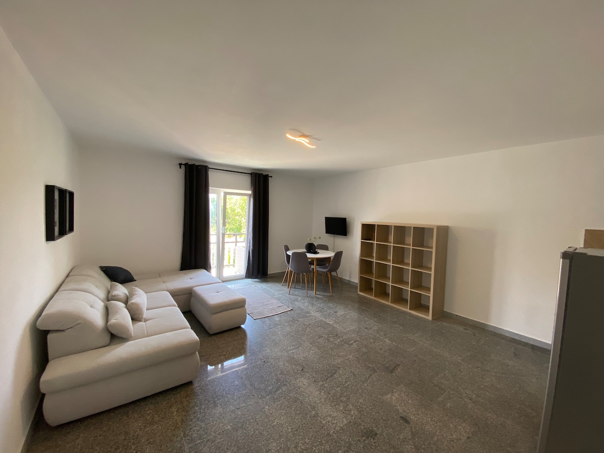 A spacious living area features a light-colored sectional sofa and a modern dining table surrounded by four chairs. Large windows provide natural light, while black curtains frame the view. A tall bookshelf occupies one corner, and a television is mounted on the wall.