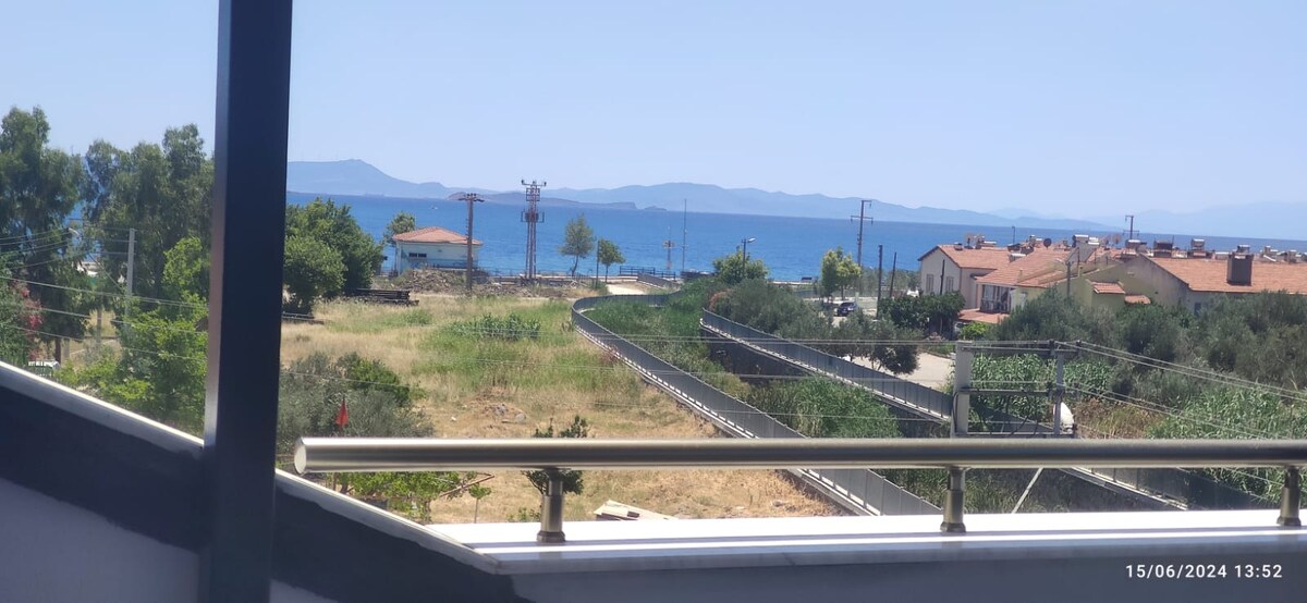 A view of the distant coastline and the sea is captured from a balcony. The landscape includes greenery and a winding road leading toward the water. Residential buildings can be seen in the background, with a clear blue sky above.