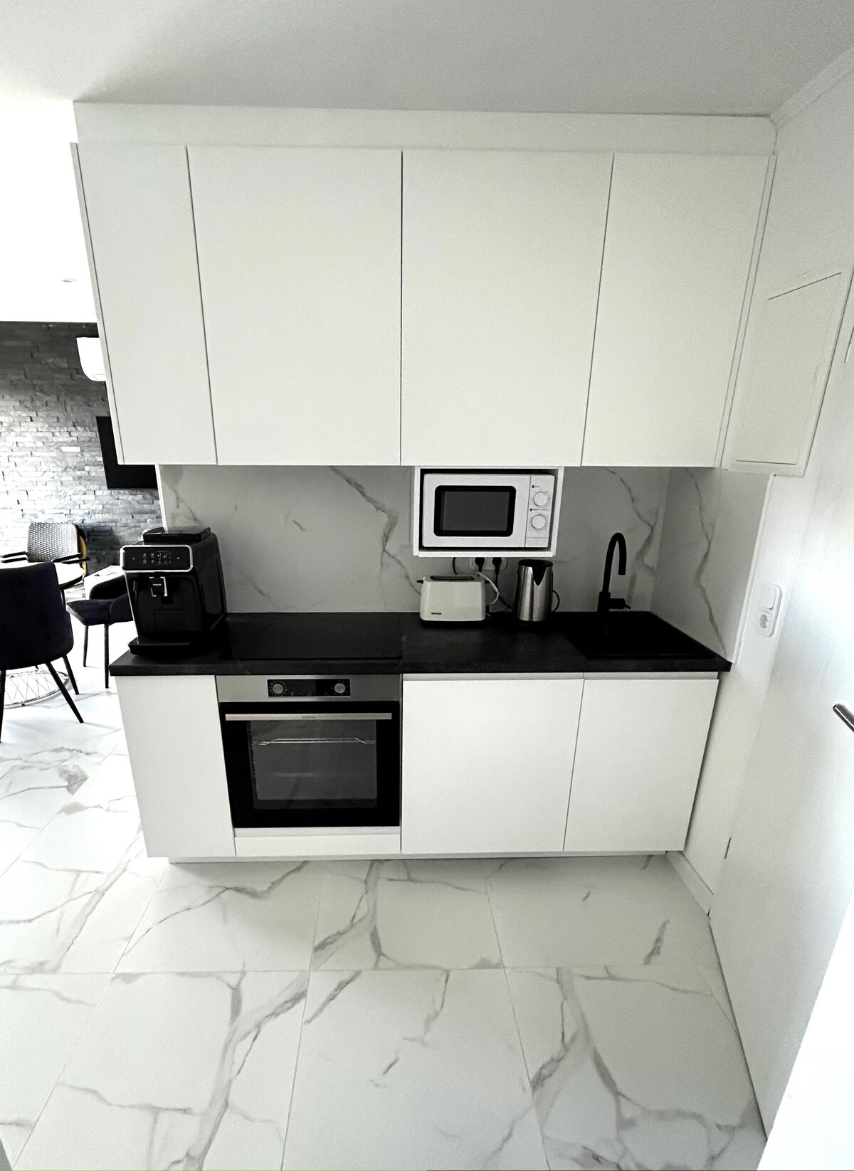 A modern kitchen area features sleek white cabinetry combined with a dark stone countertop. Included appliances are a microwave, coffee maker, and oven. The kitchen is well-lit, surrounded by marble-patterned flooring that provides a clean and elegant look.