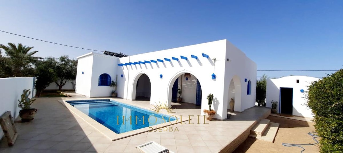 A white-walled villa is featured with blue accents, showcasing multiple arched doorways. A clear swimming pool sits in the foreground, surrounded by a tiled terrace. Lush greenery and palm trees are visible in the background, contributing to a serene outdoor setting.