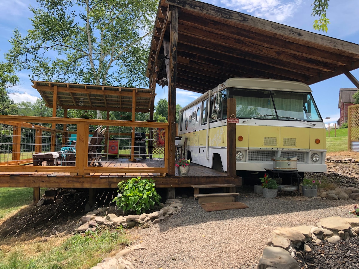 A motor home is located under a large wooden canopy on a spacious deck. Surrounding greenery includes a grassy lawn and rock borders. A shaded seating area is visible on the deck, along with potted plants that add color to the outdoor space.