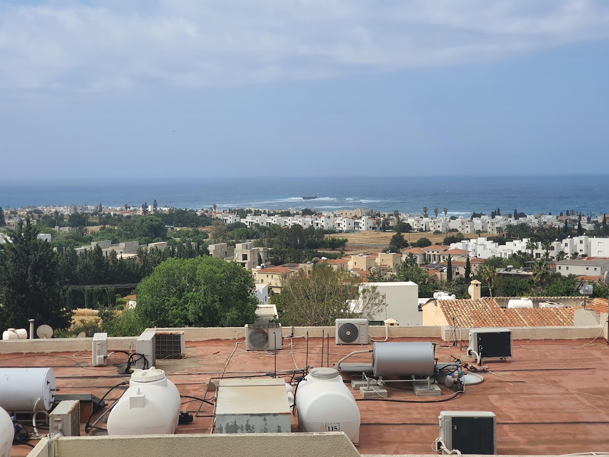 An expansive sea view is captured from the apartment balcony. The image shows rooftops and air conditioning units in the foreground, leading to a distant horizon where the sea meets the sky. Lush greenery and coastal developments are visible in the background.