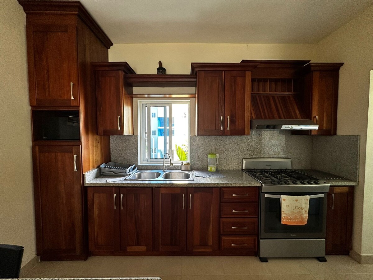 A modern kitchen is presented with wooden cabinetry and a countertop. Two sinks are located under a window that allows natural light to enter. A stainless steel gas stove and a range hood are included, providing functional space for cooking and meal preparation.