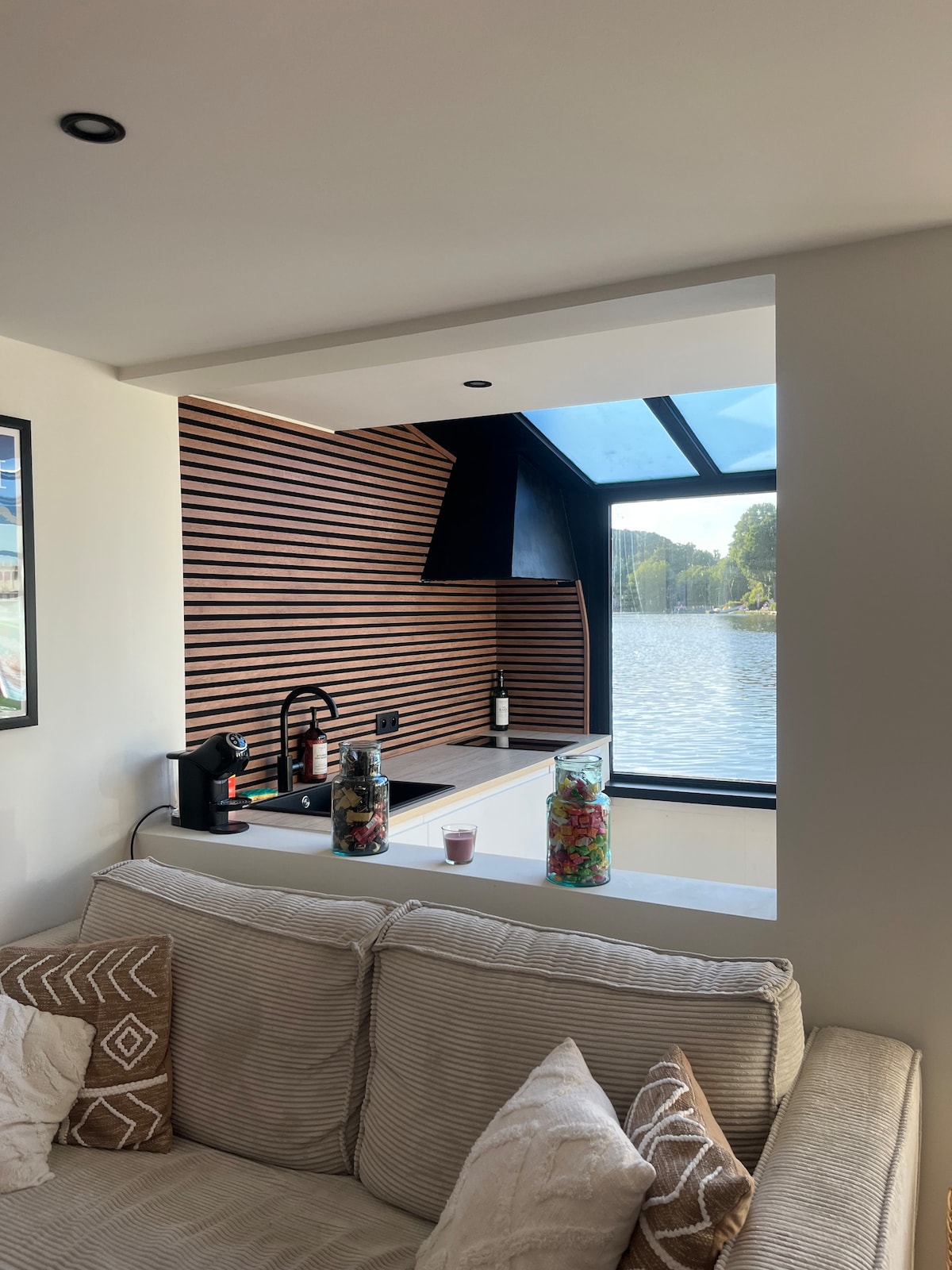 A modern kitchenette features wooden slat accents and a large window that offers a direct view of the lake. A comfortable sofa with textured cushions is positioned nearby, creating an inviting space for relaxation and connection with the serene surroundings.