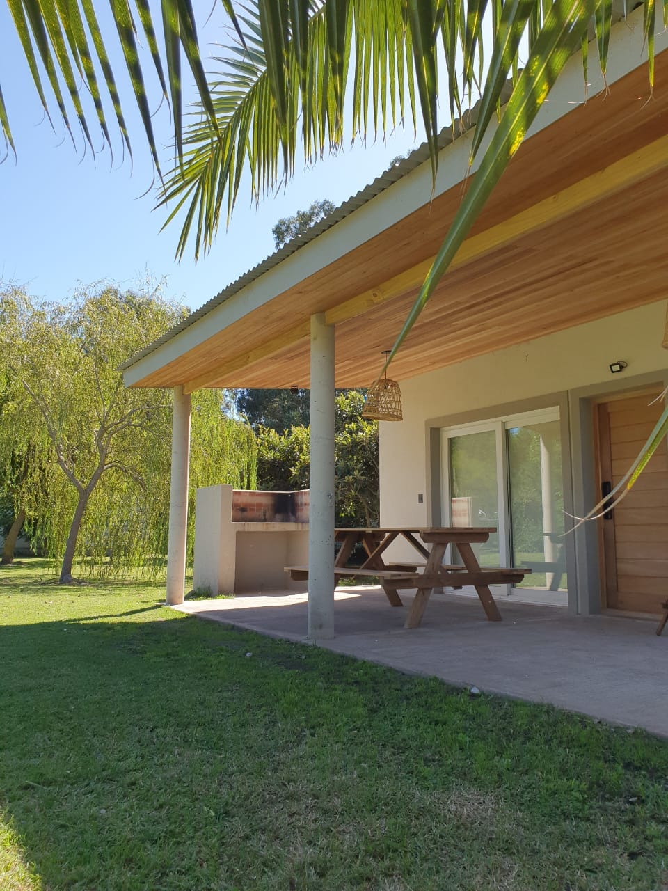 A charming outdoor space is featured, with a wooden picnic table set under a covered porch. Nearby, a built-in barbecue area is visible. Lush green grass and a young tree contribute to the serene surroundings, enhancing the connection to nature.