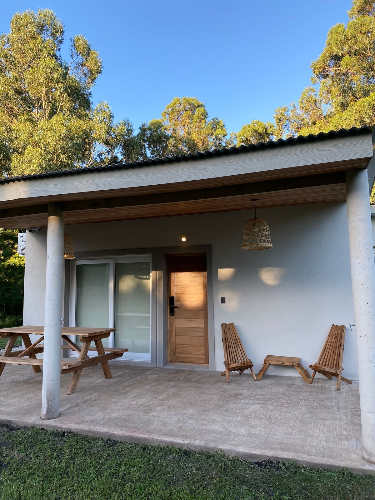 An outdoor area features a simple wooden picnic table accompanied by two lounge chairs. The entrance door displays a natural wood finish, and pendant lights hang from the ceiling. Surrounding greenery provides a serene atmosphere against the backdrop of a clear blue sky.