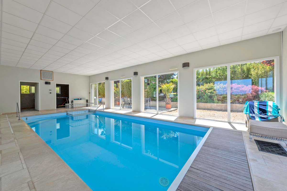 An indoor swimming pool is featured, surrounded by large glass doors that open to the outside. Natural light fills the space, illuminating the clear blue water. A seating area is visible through the doors, enhancing the functionality of the area.
