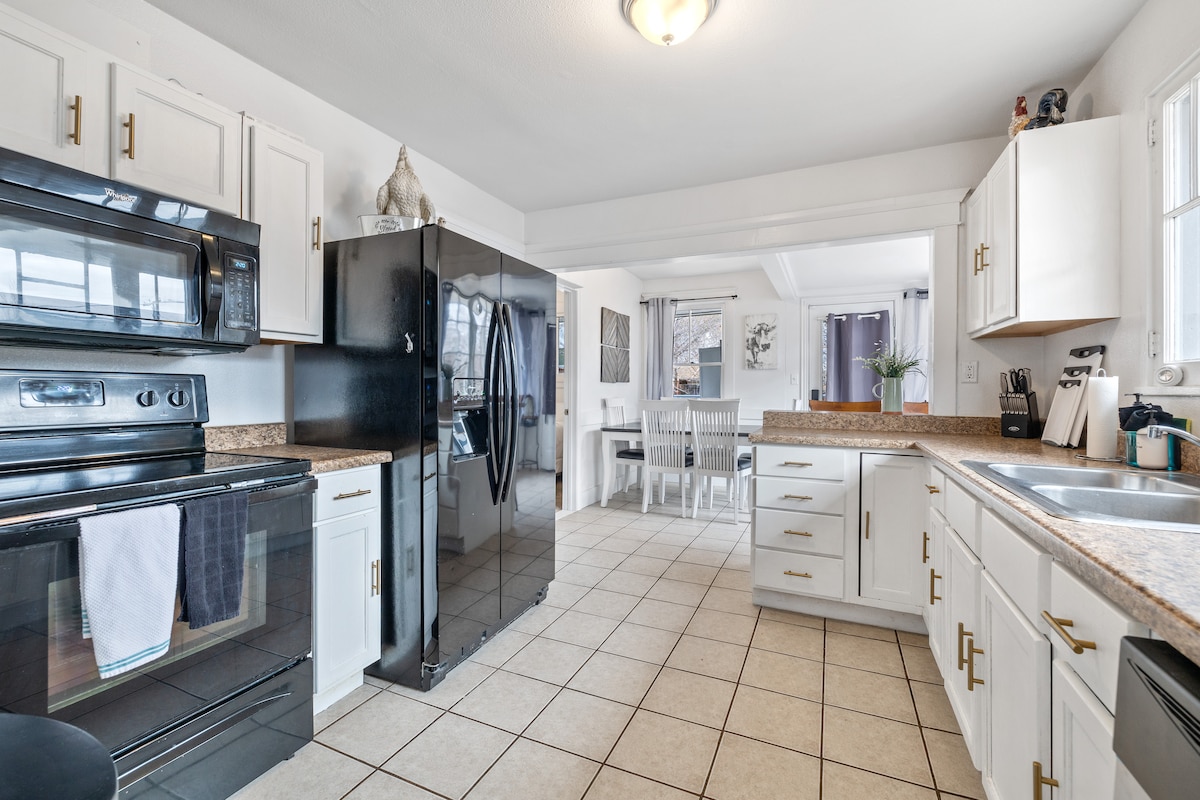The kitchen features a blend of black and white cabinetry, with a black refrigerator and stove. Spacious countertops are available for meal preparation. Natural light fills the area through multiple windows, and a dining table is visible in the adjacent dining space, fostering a functional cooking environment.