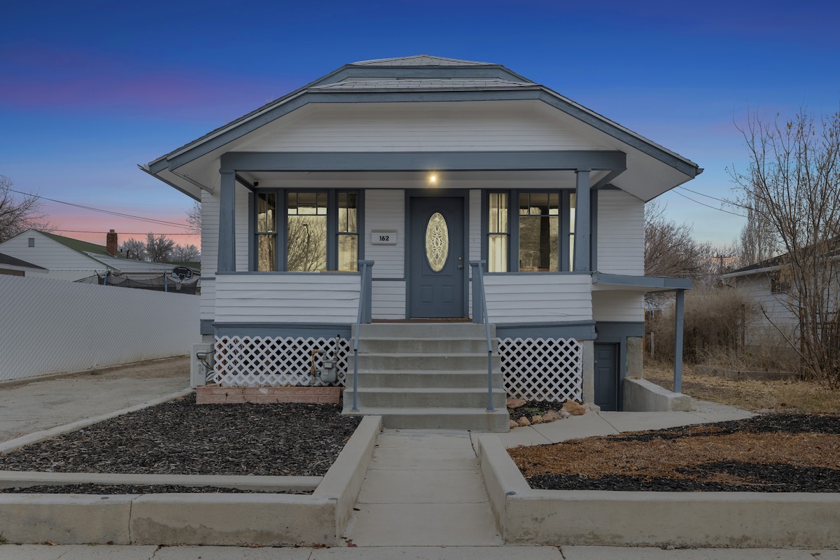The exterior of the home is presented with a welcoming front porch and white siding accented by gray trim. A set of steps leads to the main entrance, framed by large windows that allow ample natural light. Landscaping includes simple gravel and a few patches of greenery.