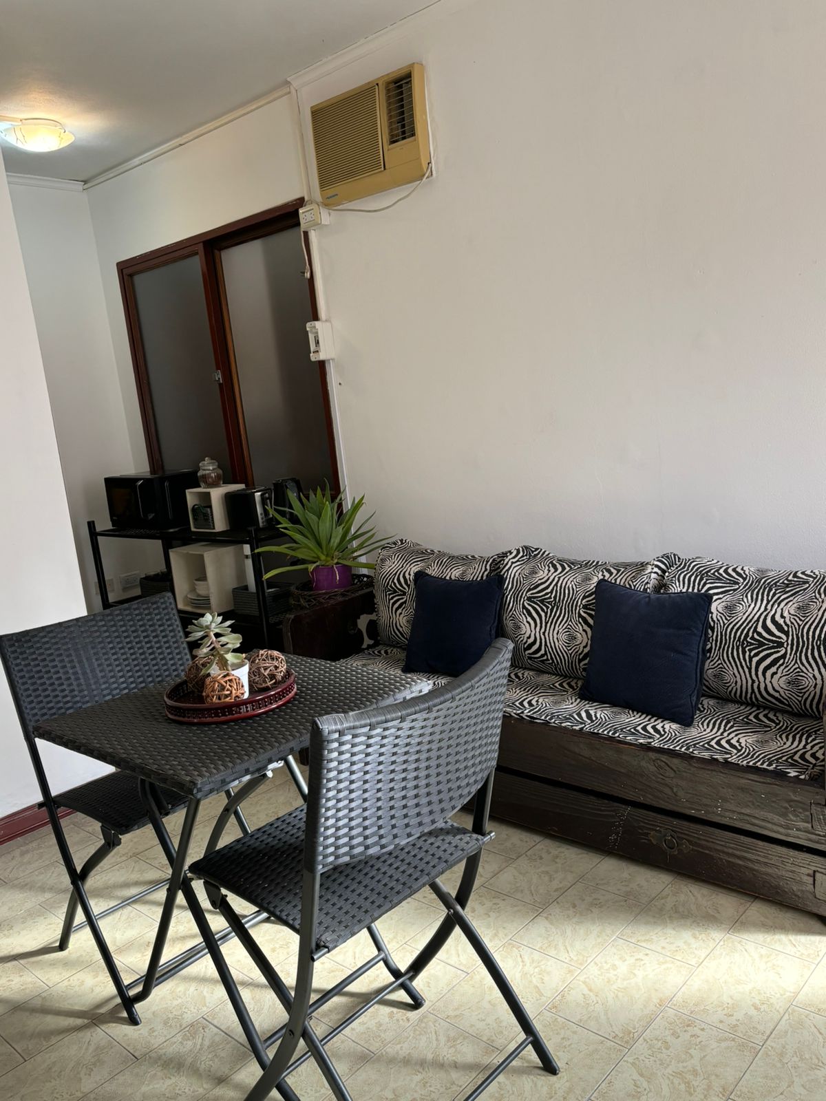 A compact dining area features a small, round table with four black chairs. A plant adds a touch of greenery, while a cozy sofa with zebra-patterned upholstery and dark cushions offers comfortable seating. A partial view of modern kitchen appliances is visible in the background.