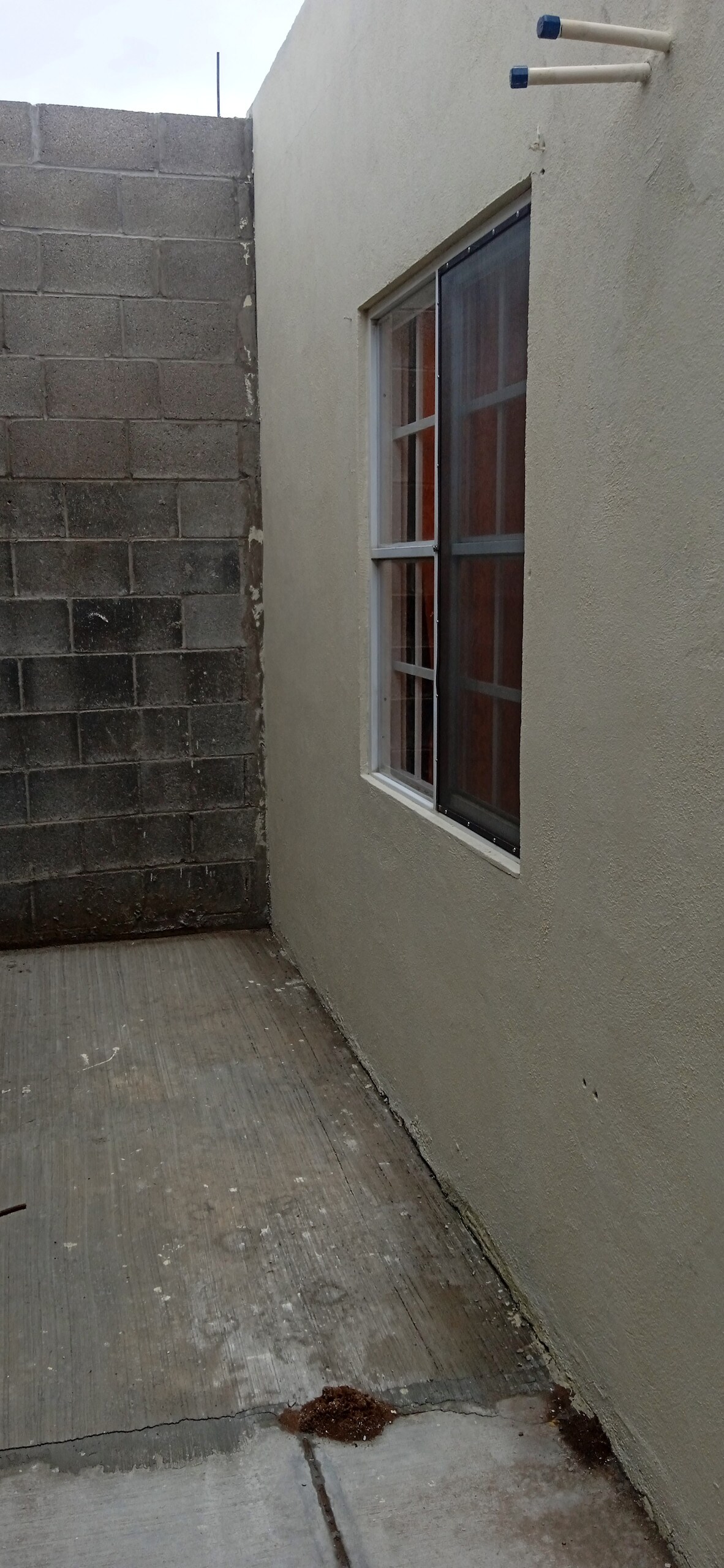 An exterior corner of a building is viewed, featuring a window with multiple panes. The surrounding walls are composed of smooth plaster and exposed concrete blocks, creating a simple and functional design. The surface below is made of concrete with visible textures.
