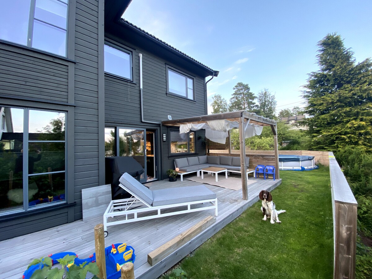 An outdoor patio area is presented with sleek lounge chairs and a shaded seating area under a pergola. A grassy space surrounds the deck, featuring a small pool in the background. A dog relaxes on the lawn, adding to the homey feel of the setting.