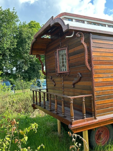 The exterior of the Pipowagen showcases finely crafted wooden paneling and elegant architectural details. A small porch with decorative railings is visible, providing a charming entrance. Surrounding greenery enhances the appeal, while the natural light highlights the rich wood tones.