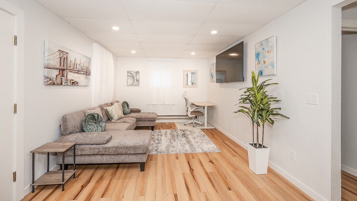 The living area features a modern design with a spacious gray sectional sofa and a stylish area rug. A flat-screen TV is mounted on the wall opposite the sitting area. Natural light is allowed in through the sheer curtains, complemented by decorative wall art and a potted plant.
