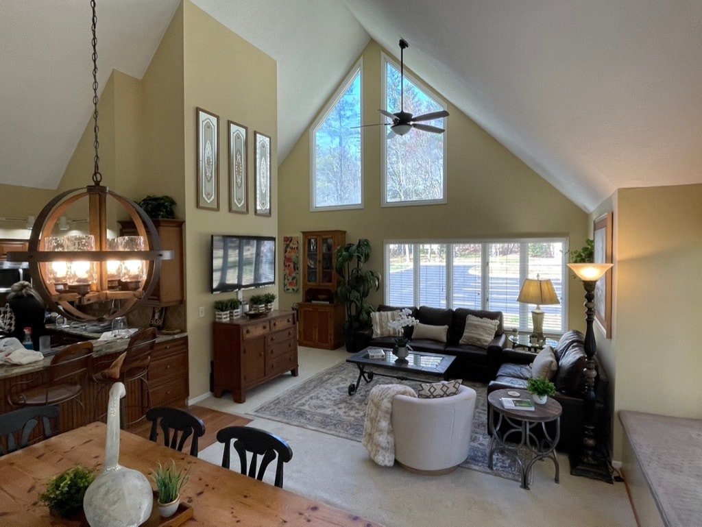 A bright living area is highlighted by tall vaulted ceilings and large windows that fill the space with natural light. Comfortable seating arrangements are present alongside a stylish dining table. Decorative elements add character, while an entertainment unit is positioned for viewing convenience.
