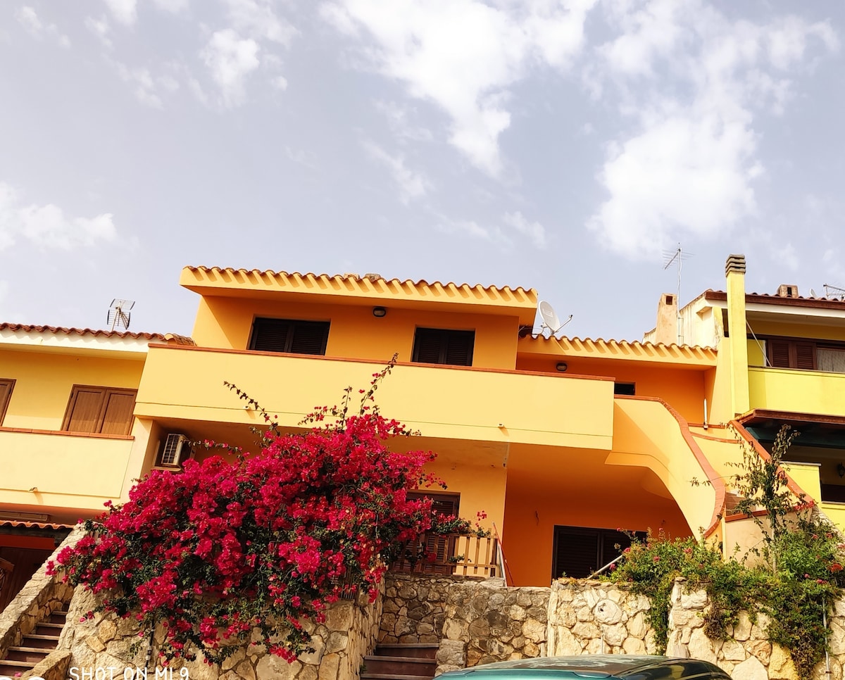 A two-story building with a vibrant yellow facade is surrounded by lush bougainvillea, showcasing vivid pink flowers. The structure features a multi-pitched roof and multiple windows, offering a cheerful appearance against a partly cloudy sky.