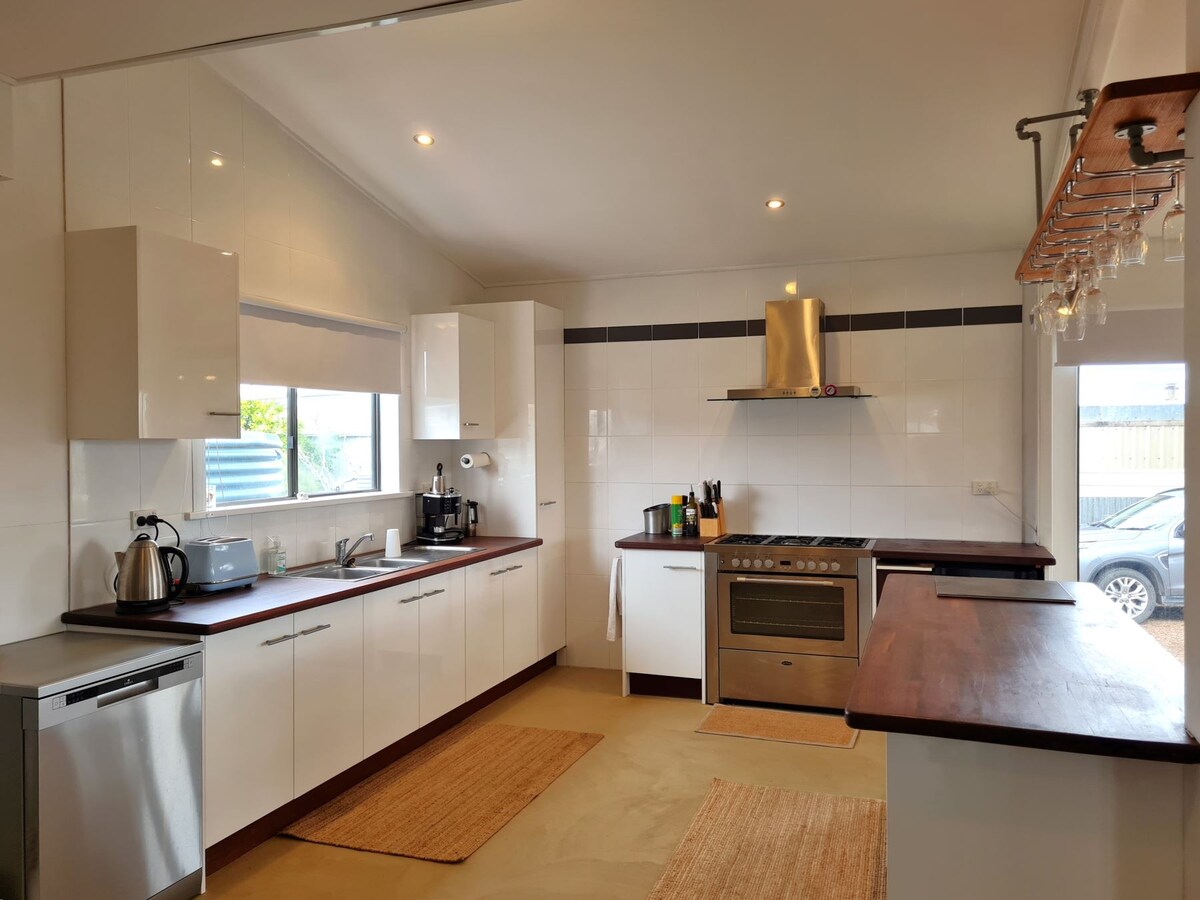 A modern kitchen is presented with sleek white cabinetry and contrasting wooden countertops. Stainless steel appliances, including a gas oven, are visible alongside a kettle and coffee machine. Natural light filters in through windows, illuminating the open space. A woven mat adds warmth to the floor.