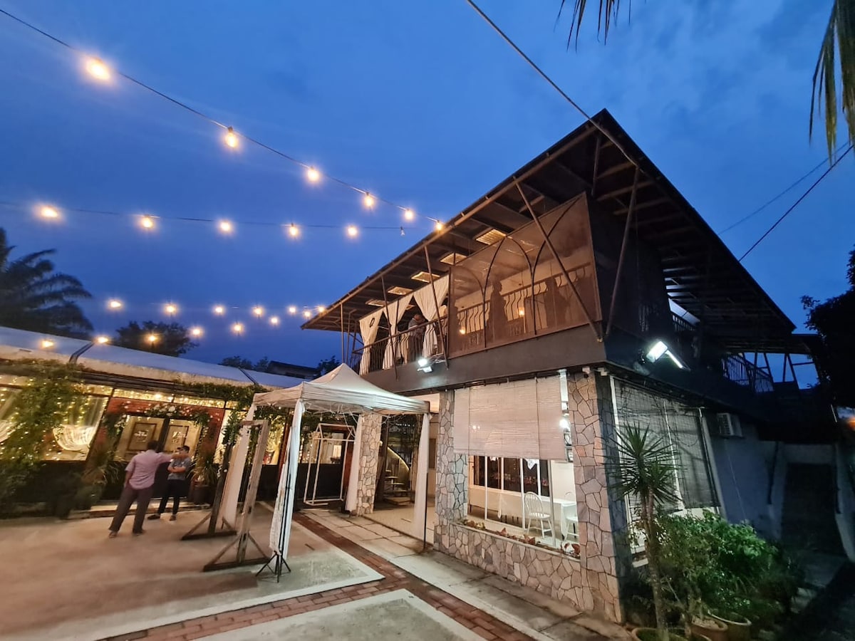An inviting bungalow exterior is illuminated by warm string lights against a dusky blue sky. The structure features a sloped roof and large windows, with a patio area visible in front. Lush greenery surrounds the space, creating a serene atmosphere.