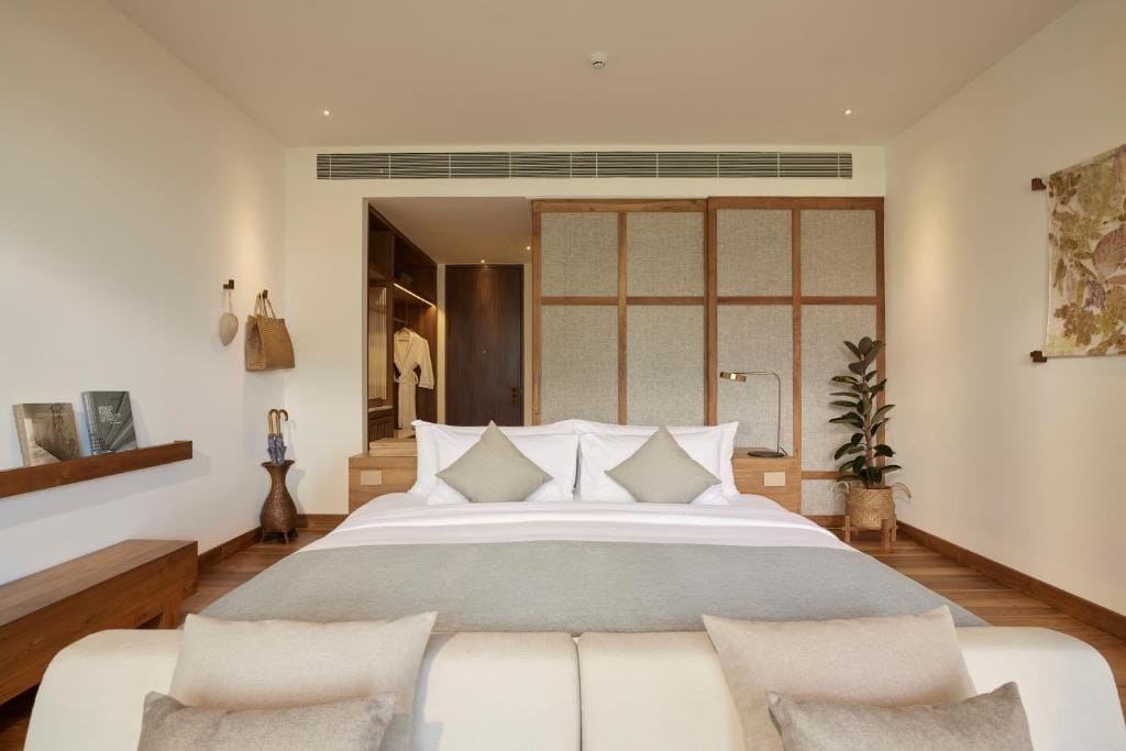 A spacious bedroom is presented with a comfortable bed dressed in neutral-toned linens. A light-colored seating area is positioned at the foot of the bed. Built-in wooden closets and a decorative plant are visible in the background, enhancing the room's calming design.